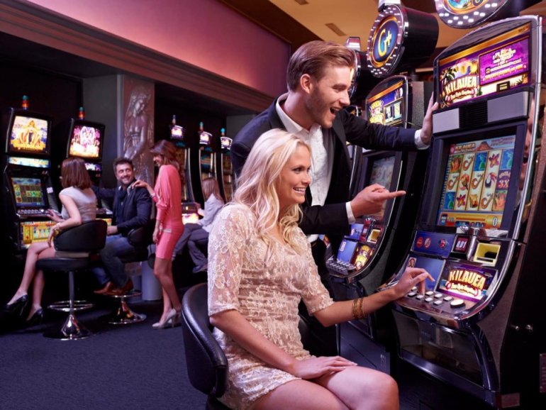 Привлекательная блондинка играет на автомате в казино, а ее спутник наблюдает за вращениями барабанов, стоя за спиной девушки